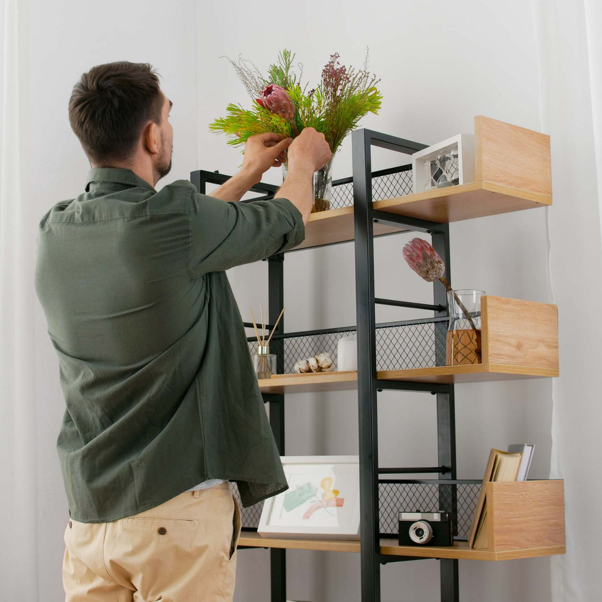 A man in an olive green shirt and beige pants is seen from behind, arranging a bouquet of green and pink flowers on a black metal and light wood floating shelf bookcase. The bookcase is styled with various home decor items against a white wall.