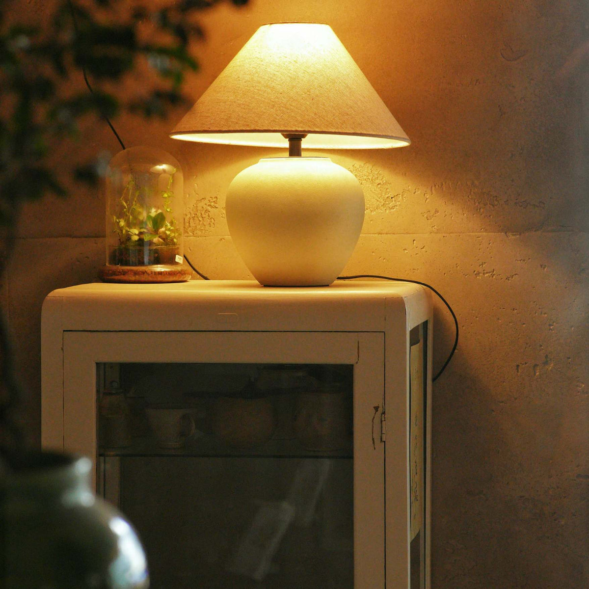 A warm white ceramic table lamp with a conical fabric shade sits lit on top of a white cabinet against a textured wall, casting a soft glow.