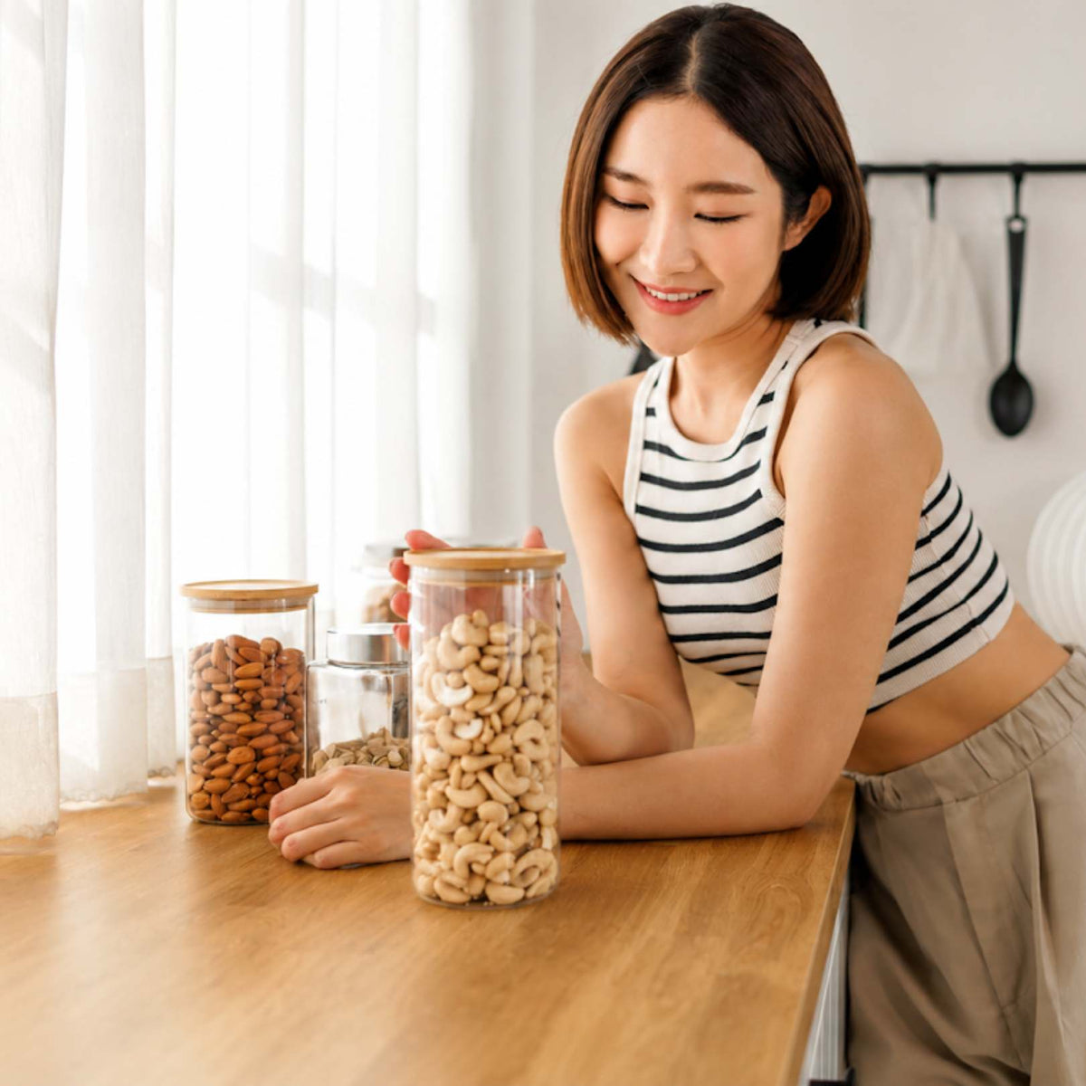 A woman in a black and white striped tank top leans on a wooden kitchen counter, smiling at clear glass canisters with wooden lids filled with nuts and dried fruits.