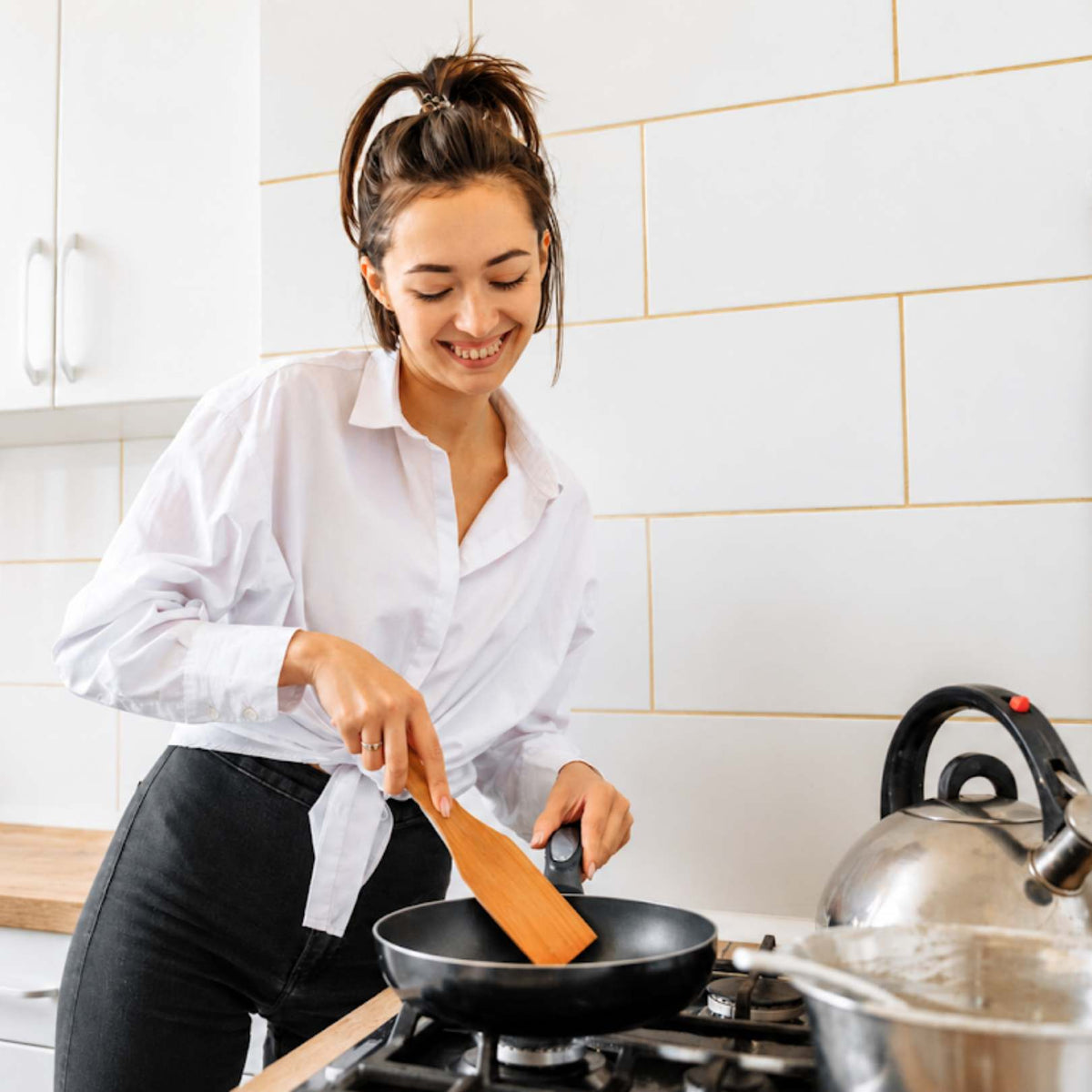 A woman in a white button-down shirt and black pants is smiling while cooking in a black frying pan on a gas stove in a bright kitchen. She is holding a wooden spatula.