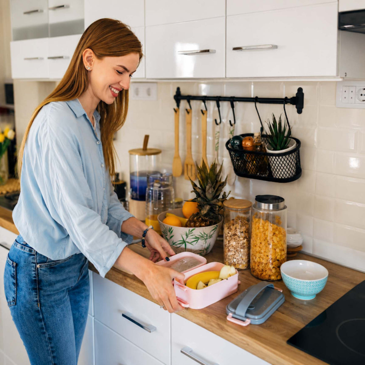 A woman in a light blue shirt and jeans is preparing a lunch box with fruit in a bright, modern kitchen. On the wooden countertop next to her are various kitchen items, including glass storage jars, a small bowl, and a gray case.