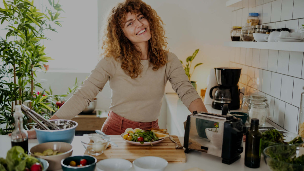 A smiling woman with curly hair stands in a bright kitchen behind a counter filled with food prep items, bowls, a juicer, and plants.