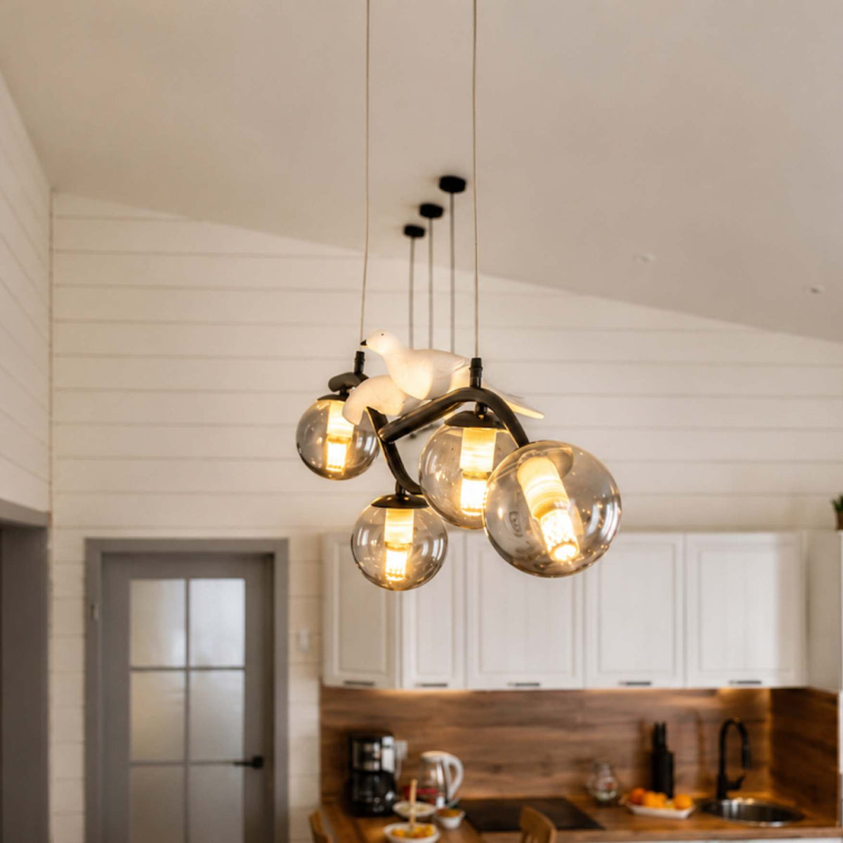 A modern multi-light pendant chandelier with four clear glass globe shades and a white dove-shaped light fixture, hanging in a bright kitchen with white shiplap walls and white cabinets.
