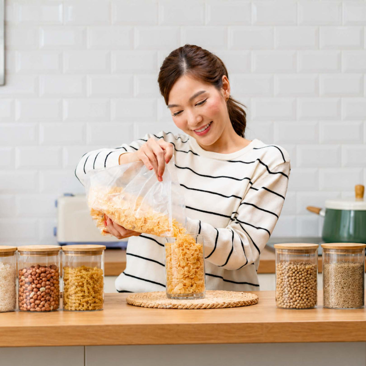 A woman in a kitchen pours dry pasta from a clear plastic bag into a glass jar. Several clear storage jars filled with dried grains and pasta are arranged on a wooden countertop against a white brick wall background.
