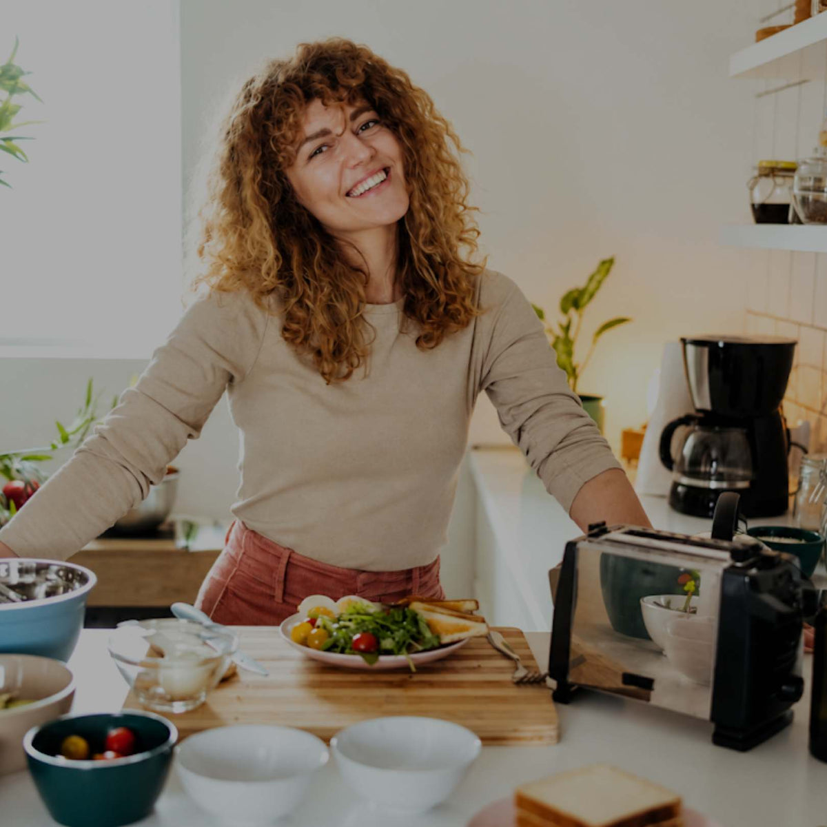A smiling woman with curly hair stands in a bright kitchen behind a counter with food, bowls, and kitchen appliances. She is wearing a beige long-sleeve top and pink pants.
