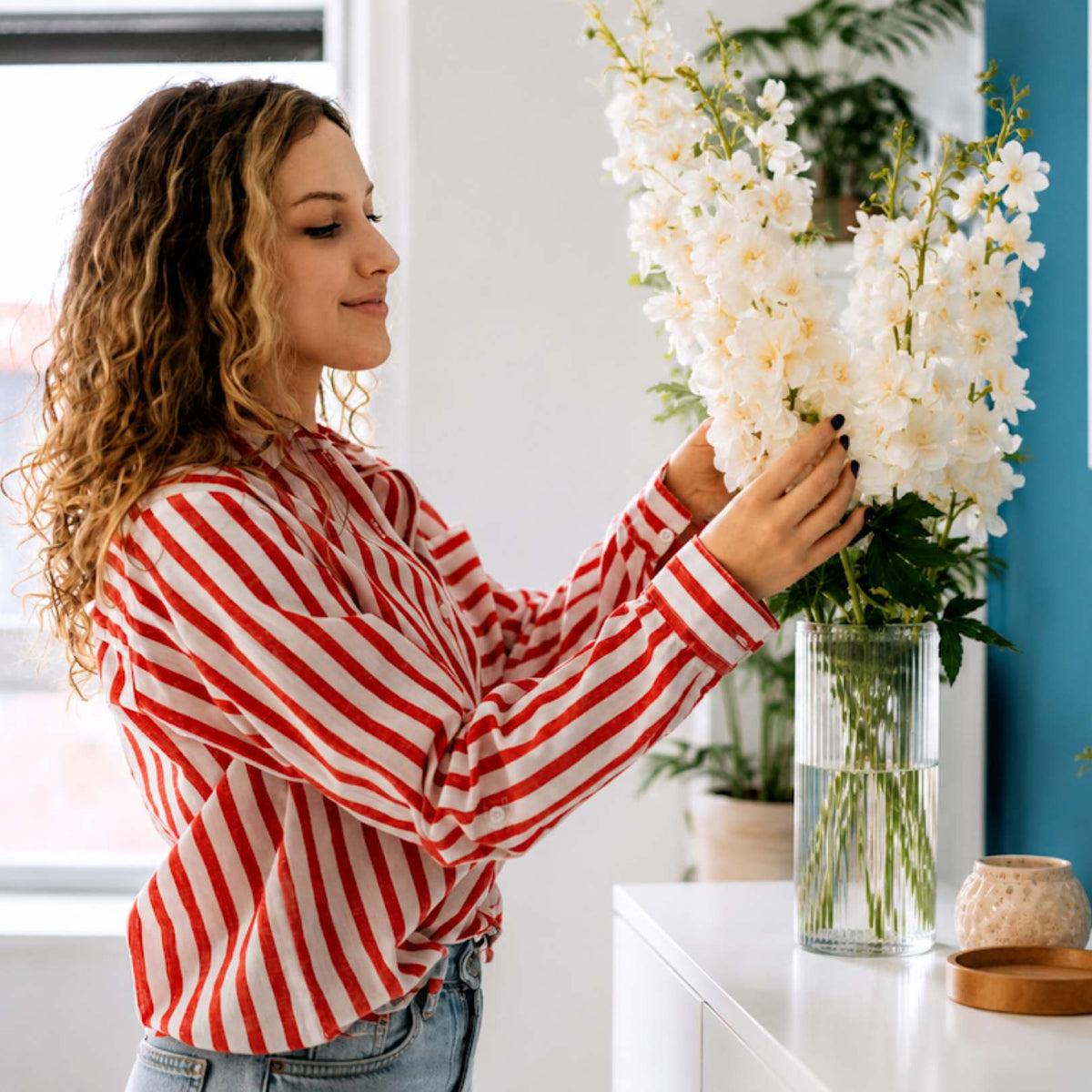 A woman with curly brown hair wearing a red and white striped long-sleeve shirt and blue jeans is holding a large bouquet of white flowers in a clear glass vase on a white table.